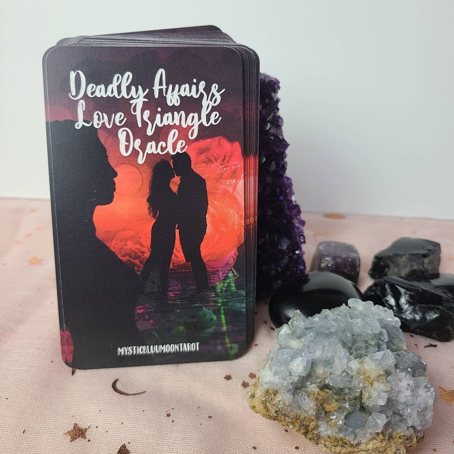 May include: A deck of oracle cards with a black background and colorful imagery. The title of the deck is "Deadly Affairs Love Triangle Oracle" with the text "MysticellumoonTarot" at the bottom.